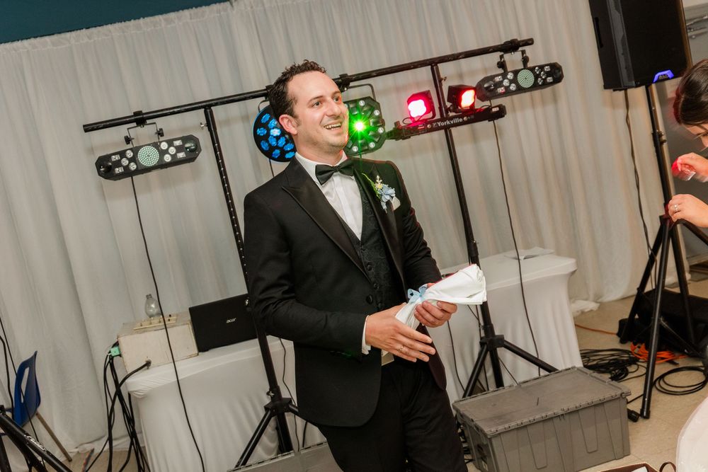 Groom in black tuxedo smiling during wedding reception with colorful DJ lights in background.