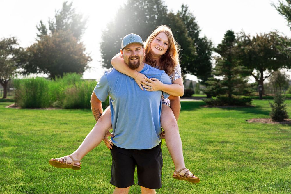 A smiling man gives a laughing woman a piggyback ride on a sunny green lawn with trees in the background.