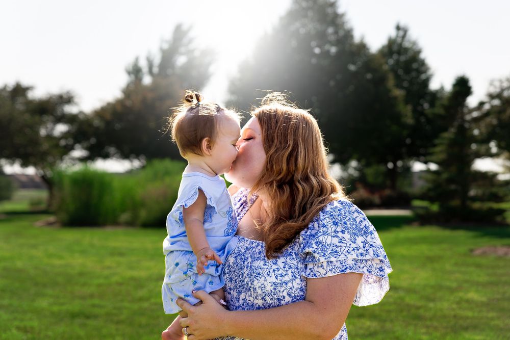 A mother in a blue floral dress kisses her baby girl outdoors in a sunny green park.