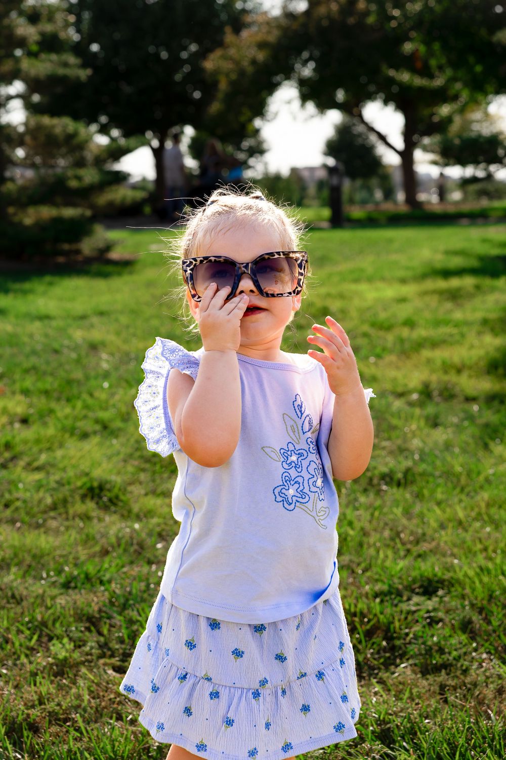 Cute toddler girl wearing oversized leopard sunglasses and blue floral outfit standing on green grass in a park.