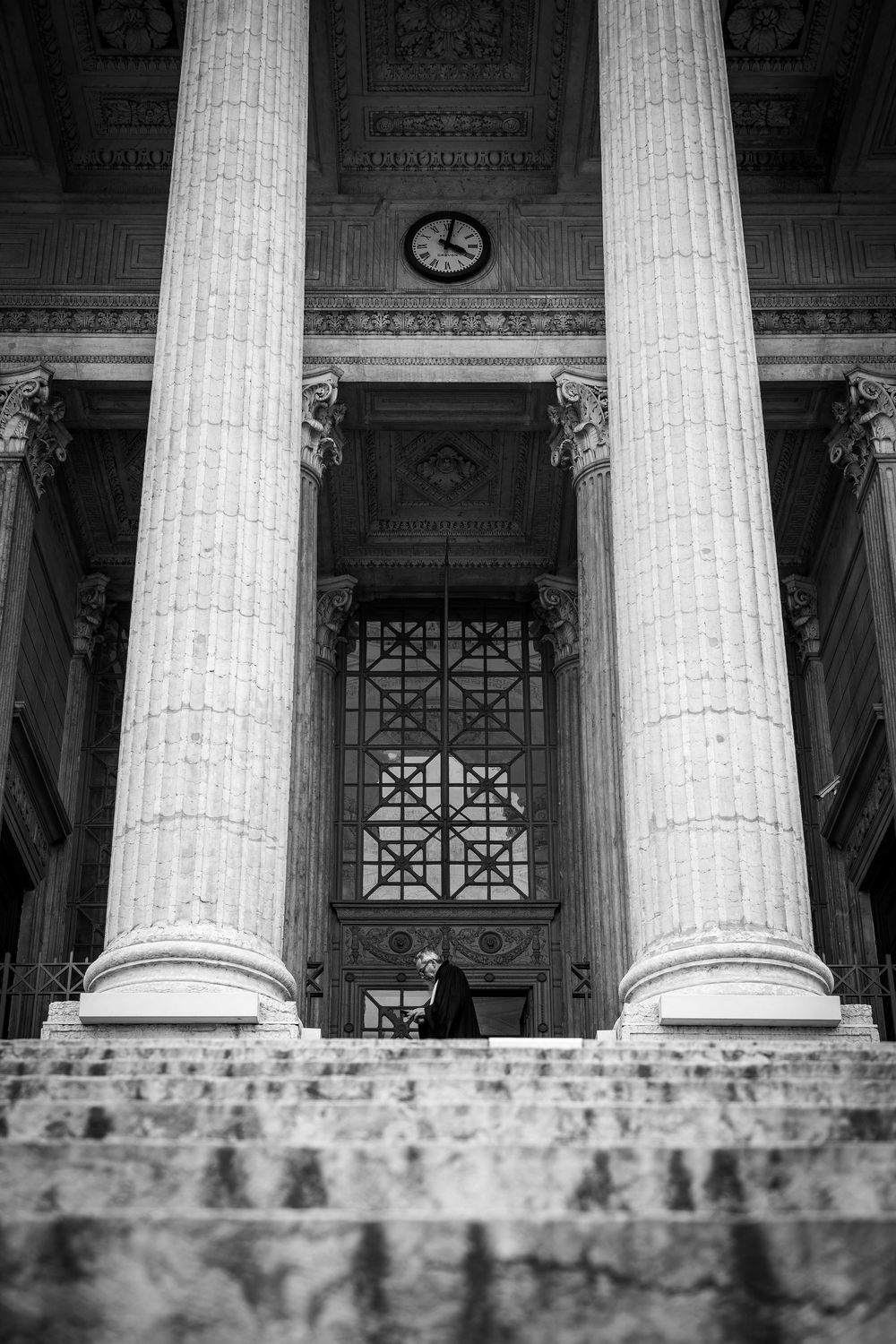 Passant devant les colonnes du Palais de Justice de Lyon, photographié en noir et blanc et en contre-plongée.