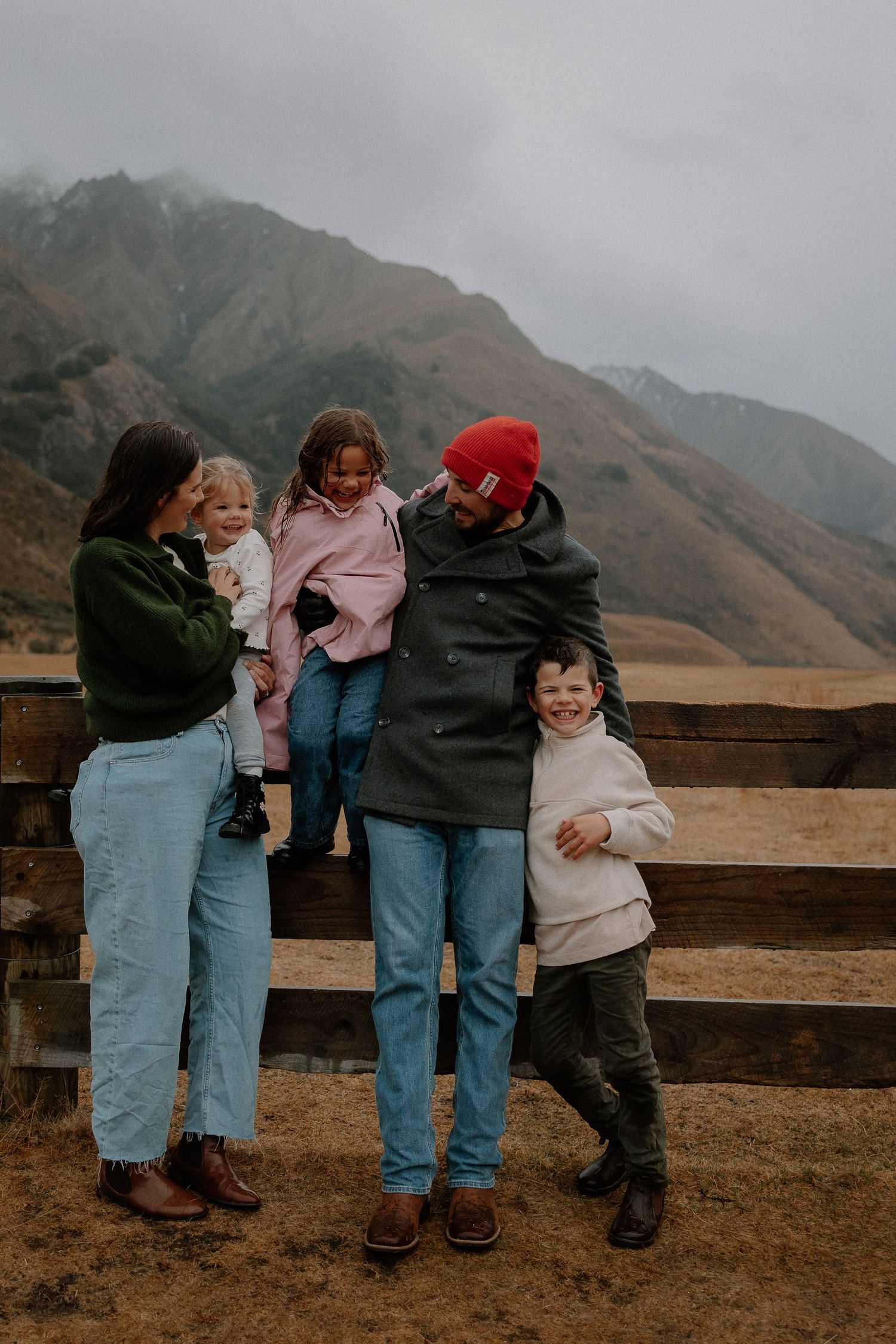 A woman playfully holds a laughing child upside down in a scenic mountain field on an overcast day.