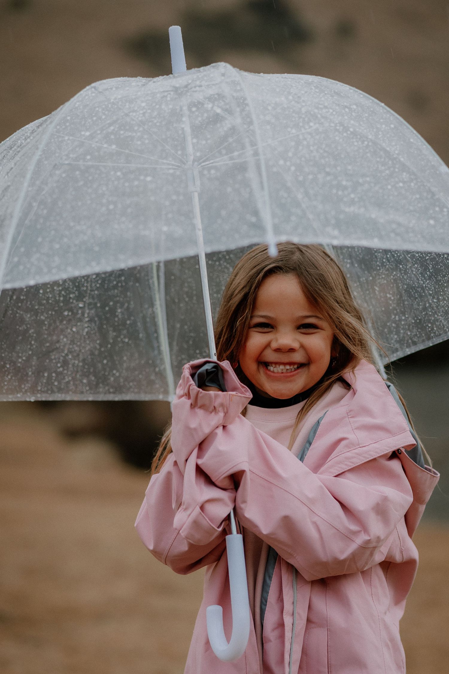 Smiling young girl in pink raincoat holding a clear umbrella outdoors in the rain.