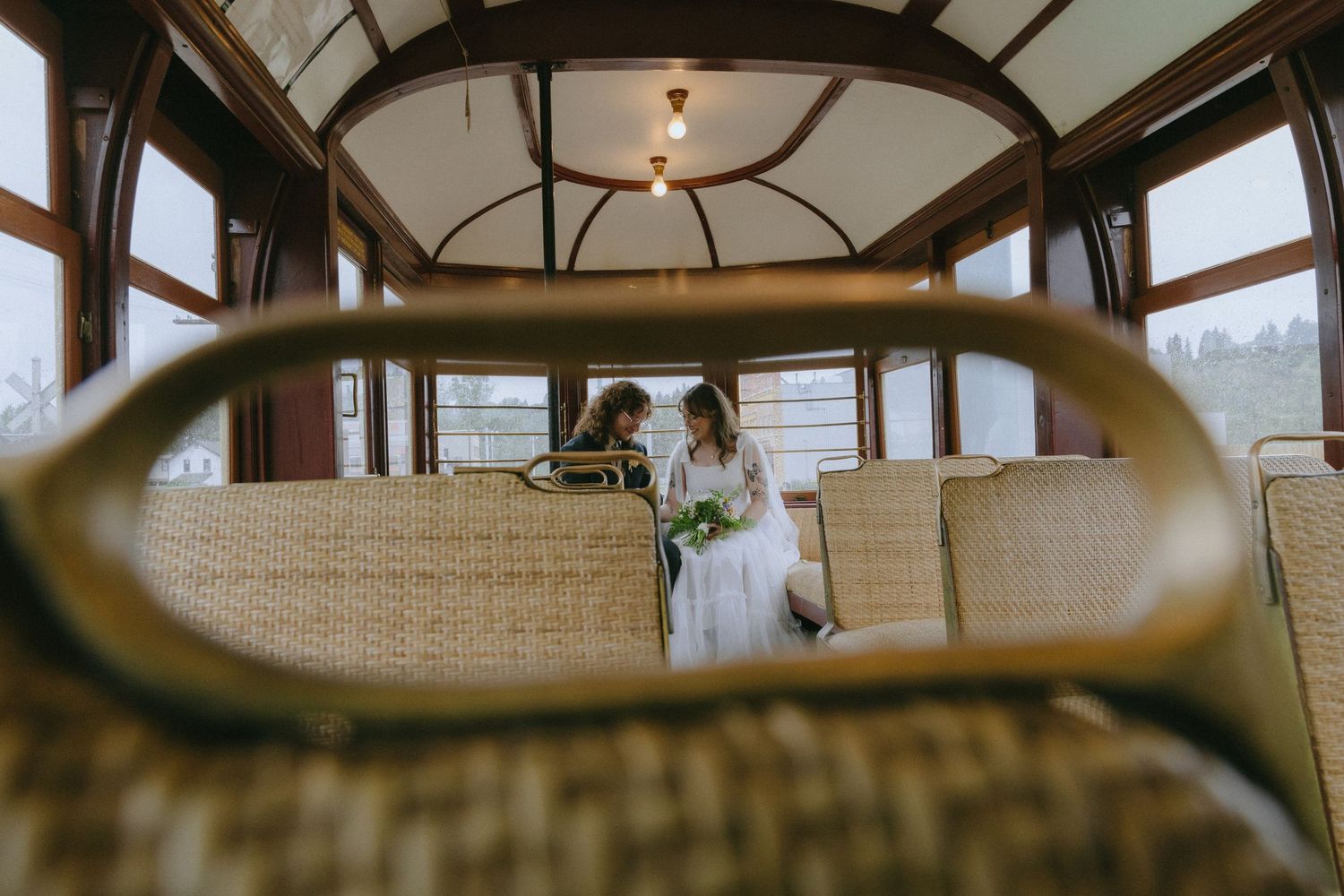 Bride in white gown holding bouquet inside vintage trolley car with wicker seats and warm wood interior.