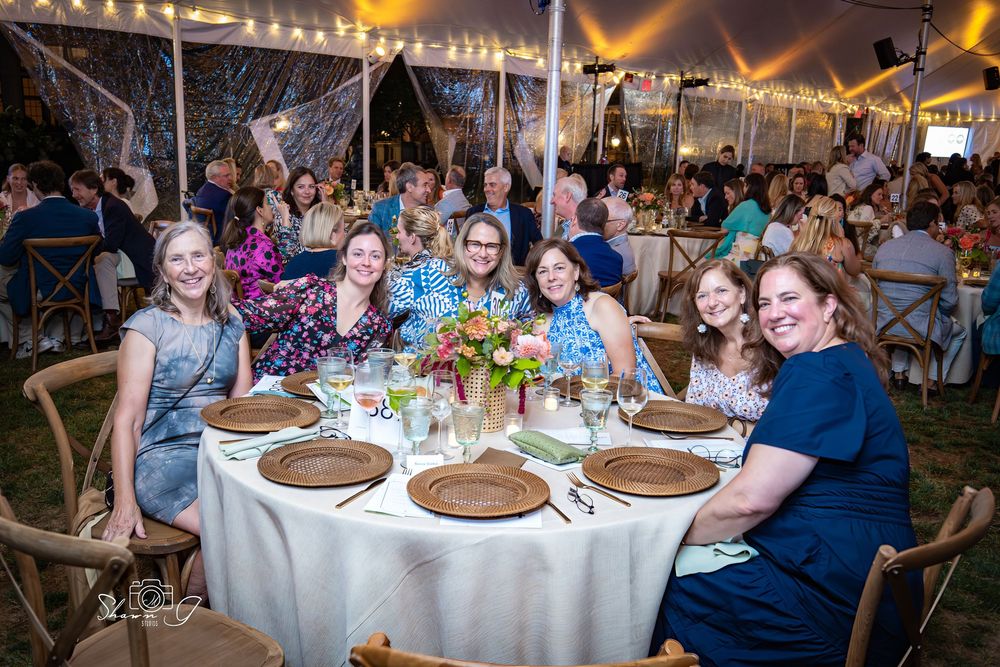 Women smiling and posing together at a festive outdoor tent gala dinner with floral centerpieces and string lights.
