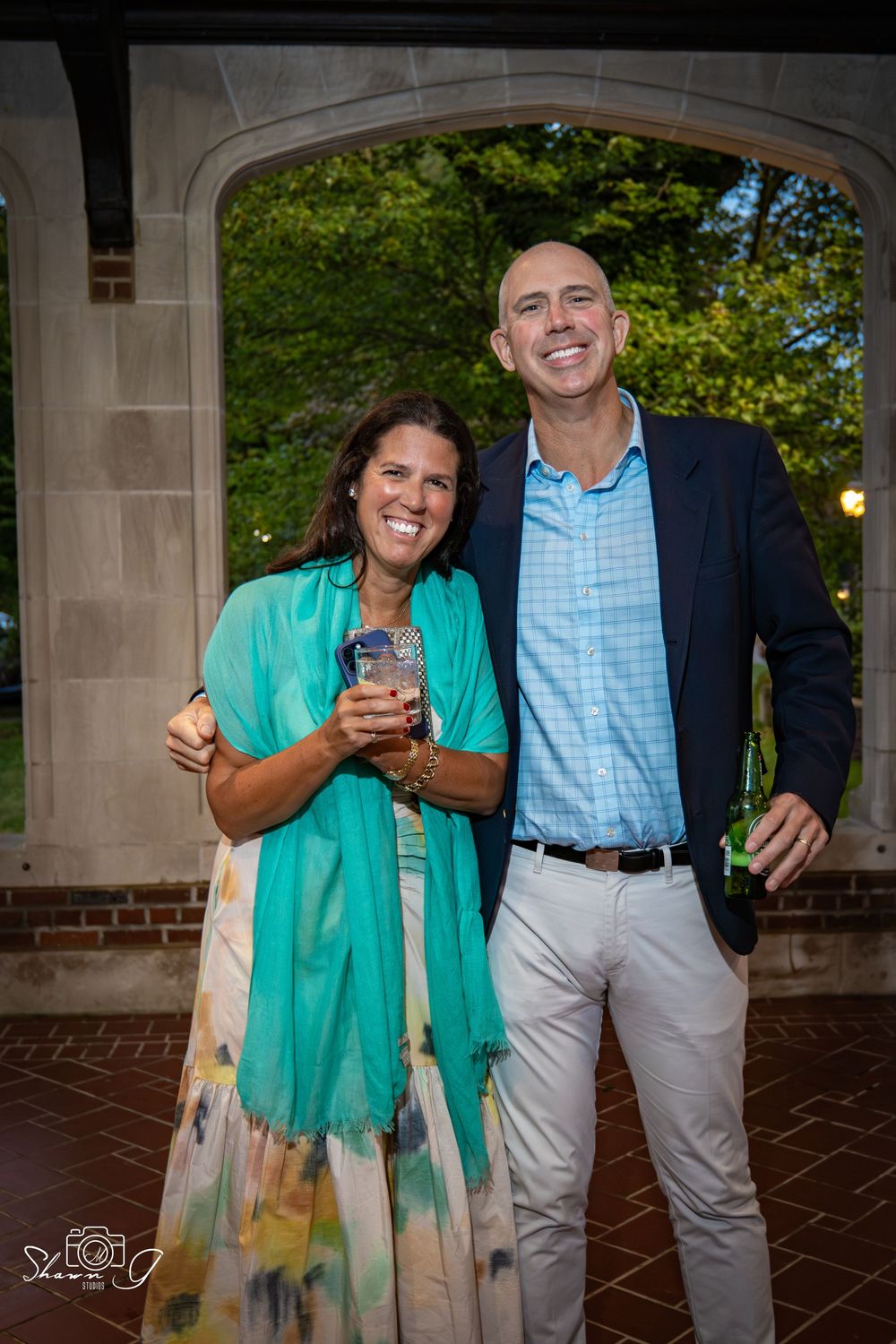 A smiling couple poses outdoors at an evening event, woman in teal wrap dress, man in blue blazer and khakis.