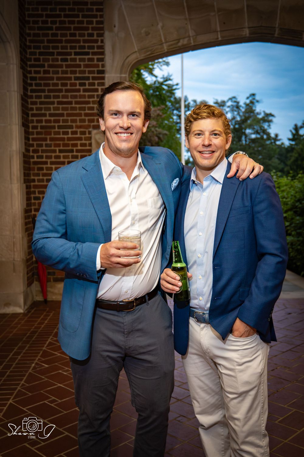 Two smiling men in blue blazers holding drinks pose together under a brick archway at an outdoor event.