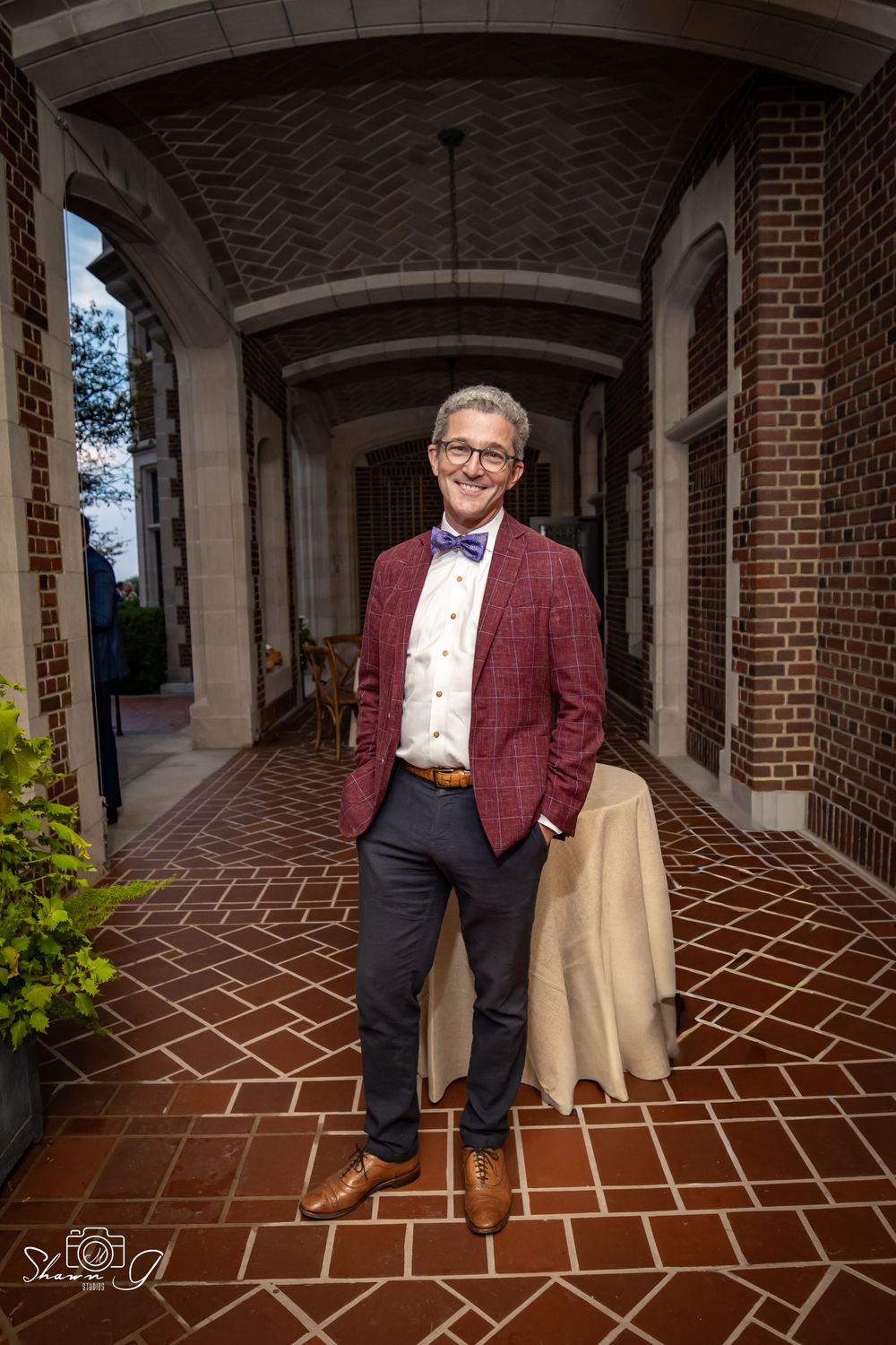 Man in burgundy blazer and bow tie smiling in brick archway corridor.