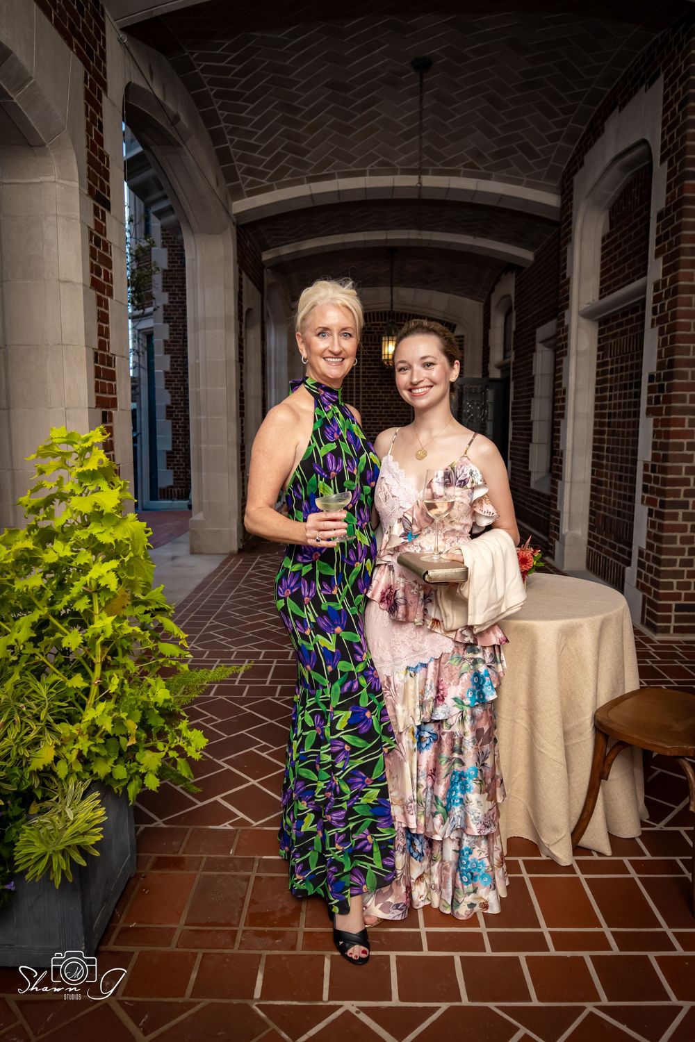 Two elegantly dressed women pose together in a brick archway corridor at a formal event.