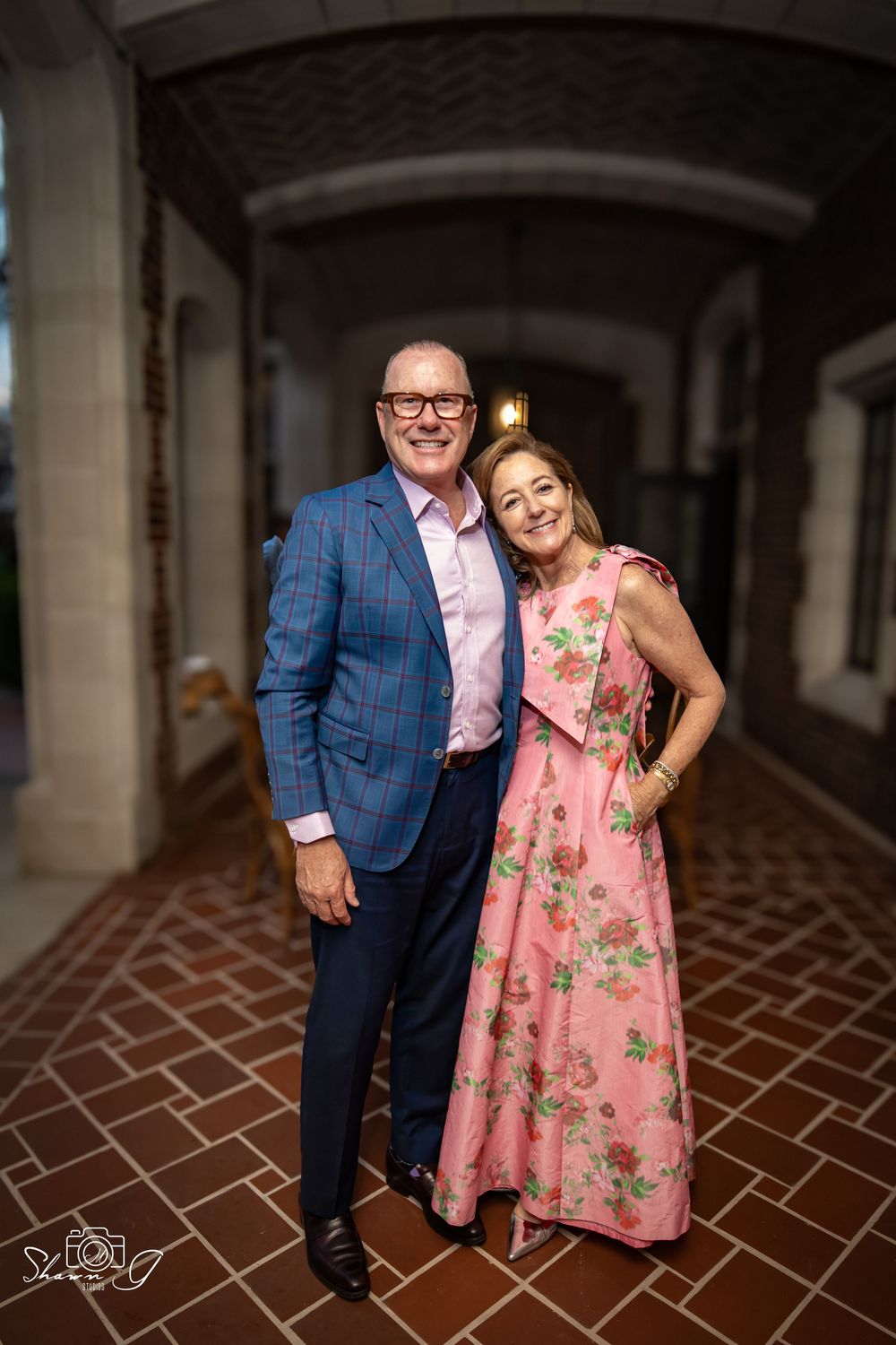 Smiling couple posing in elegant attire, man in blue plaid blazer and woman in pink floral dress, standing in brick archway.