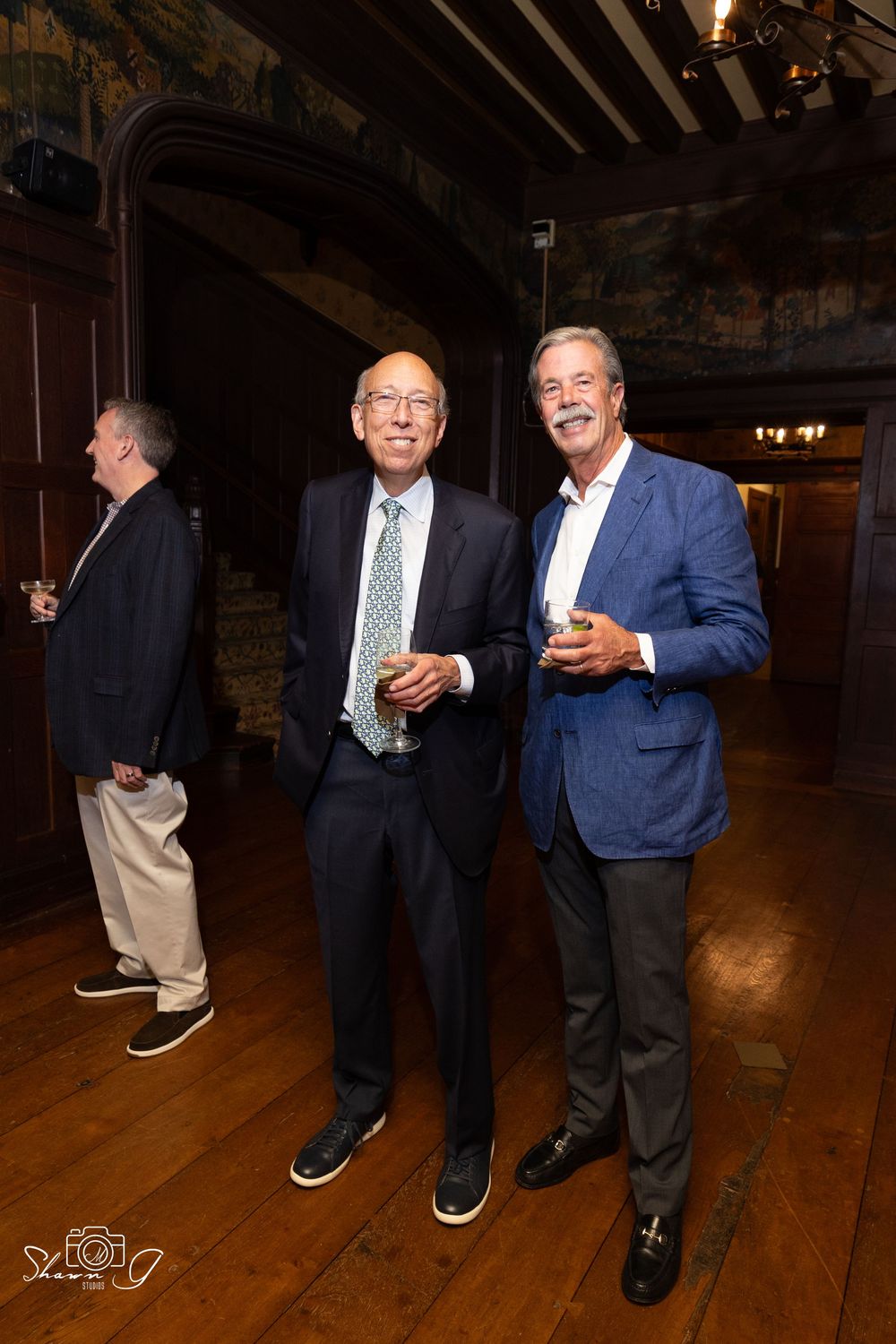 Two well-dressed men in suits smiling and holding drinks at an elegant wood-paneled event venue.