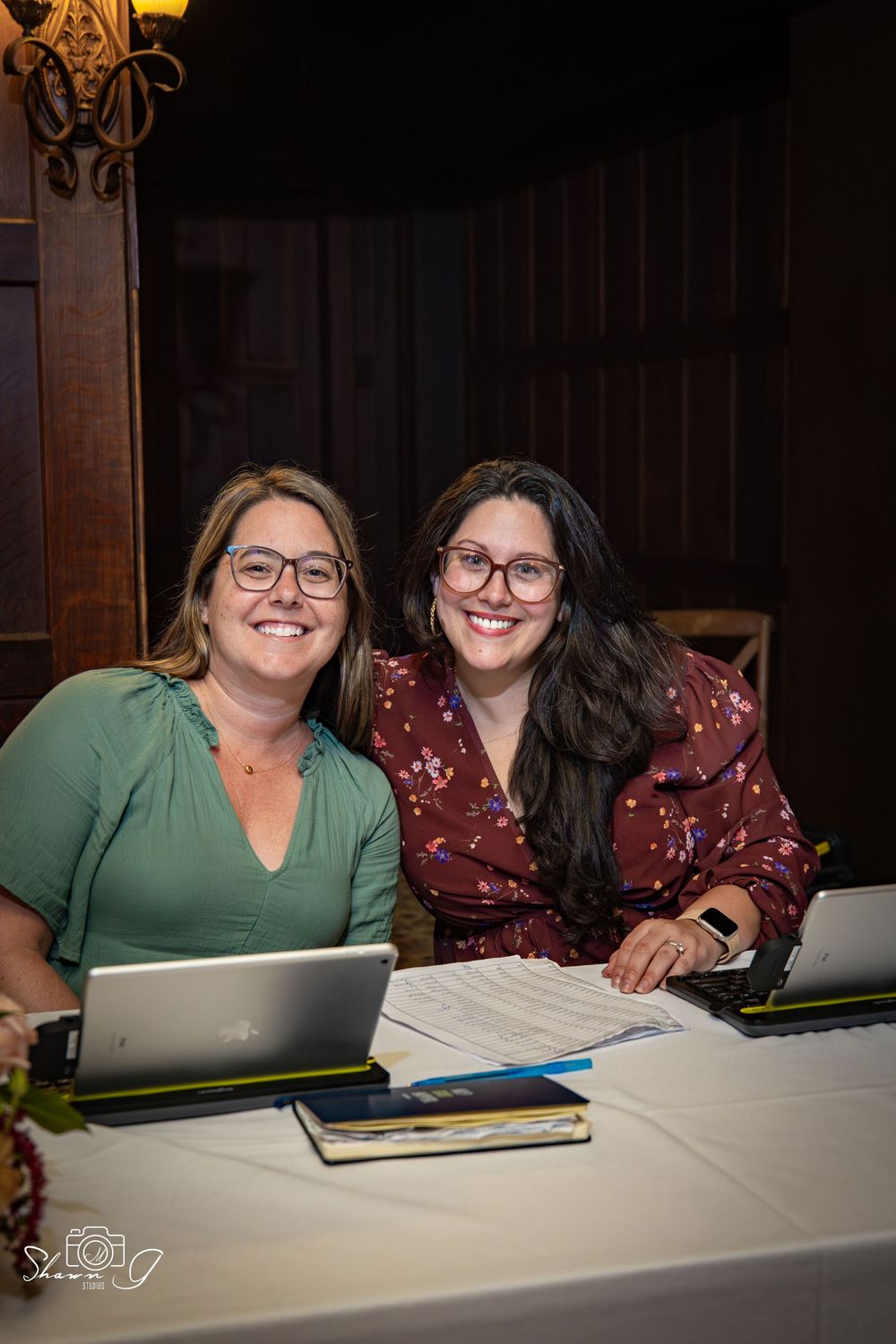 Two smiling women wearing glasses sit together at a table with laptops and documents in a warmly lit room.
