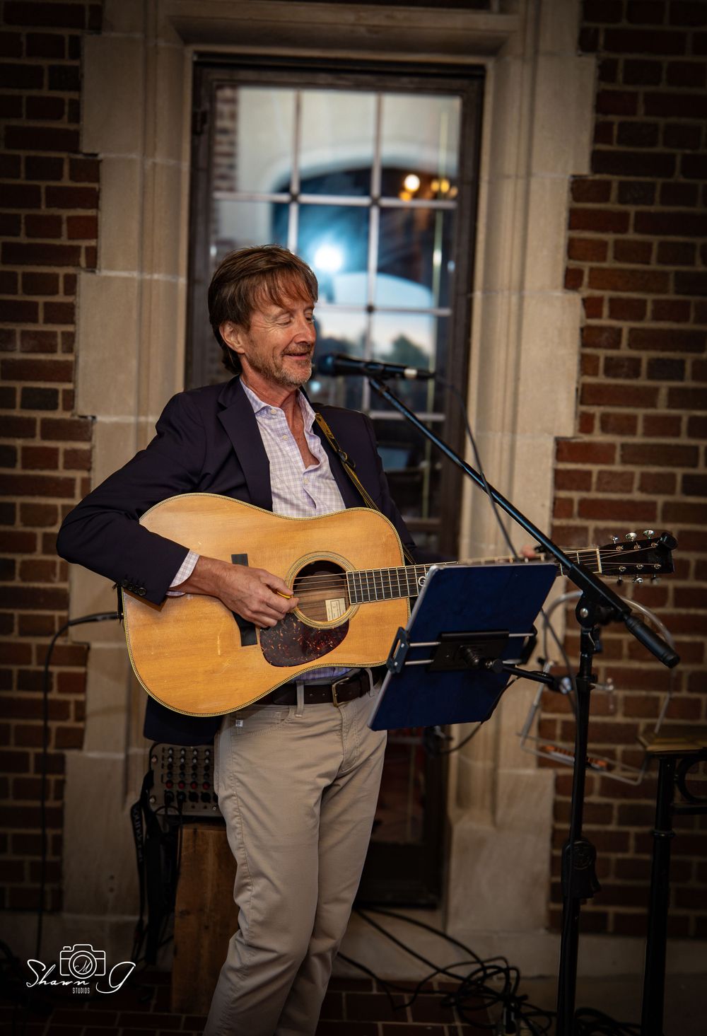 Man playing acoustic guitar at microphone stand indoors, smiling, wearing blazer, brick wall background.