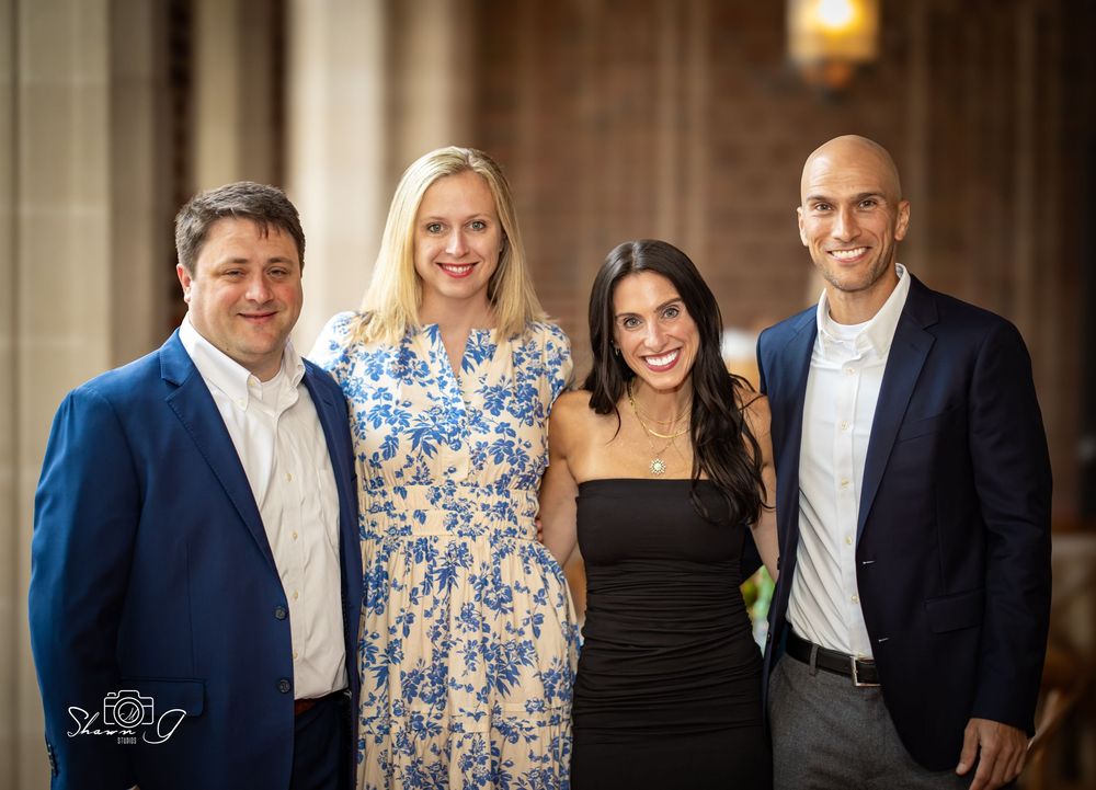 Four smiling adults posing together at a formal event, dressed in elegant attire including a blue suit and floral dress.