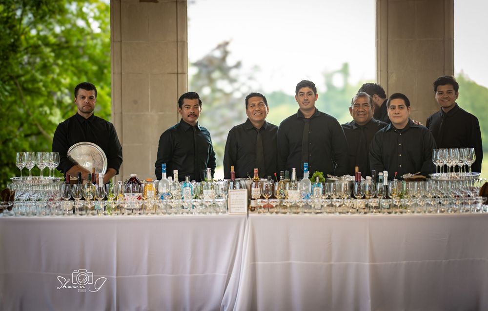 Catering staff in black uniforms stand behind a banquet bar table with glassware and bottles at an outdoor event.