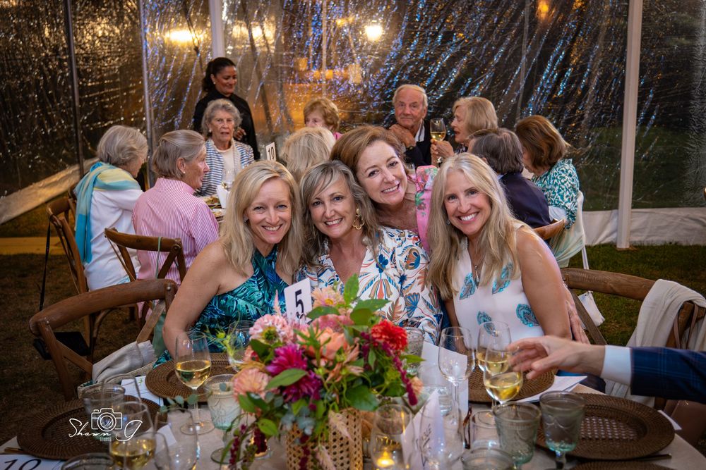 Four women smile together at an elegant outdoor evening event under a tent with string lights and floral centerpieces.