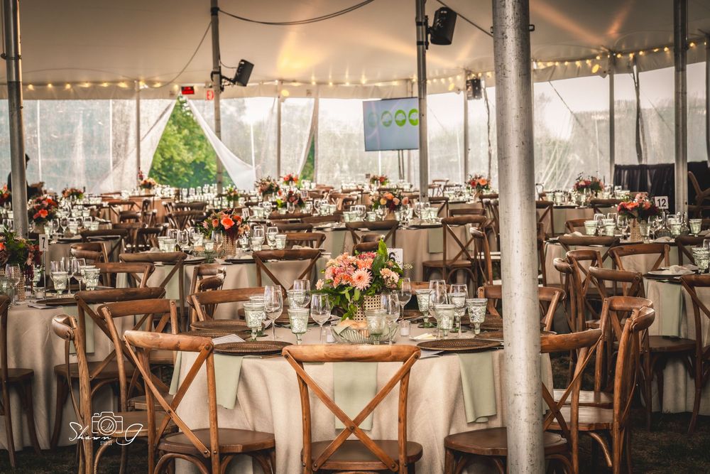 Elegant outdoor wedding reception tent with round tables, wooden cross-back chairs, floral centerpieces, and string lights.