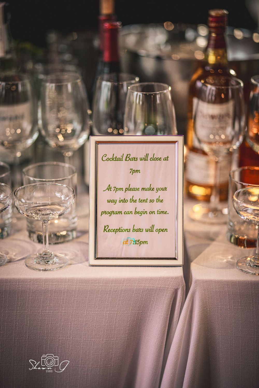 Elegant wedding reception table with framed cocktail bar schedule sign, wine glasses, and liquor bottles on white linen.