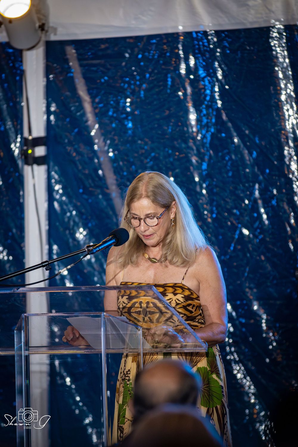 Woman in patterned dress speaking at a clear podium with microphone at a formal event with blue sequin backdrop.