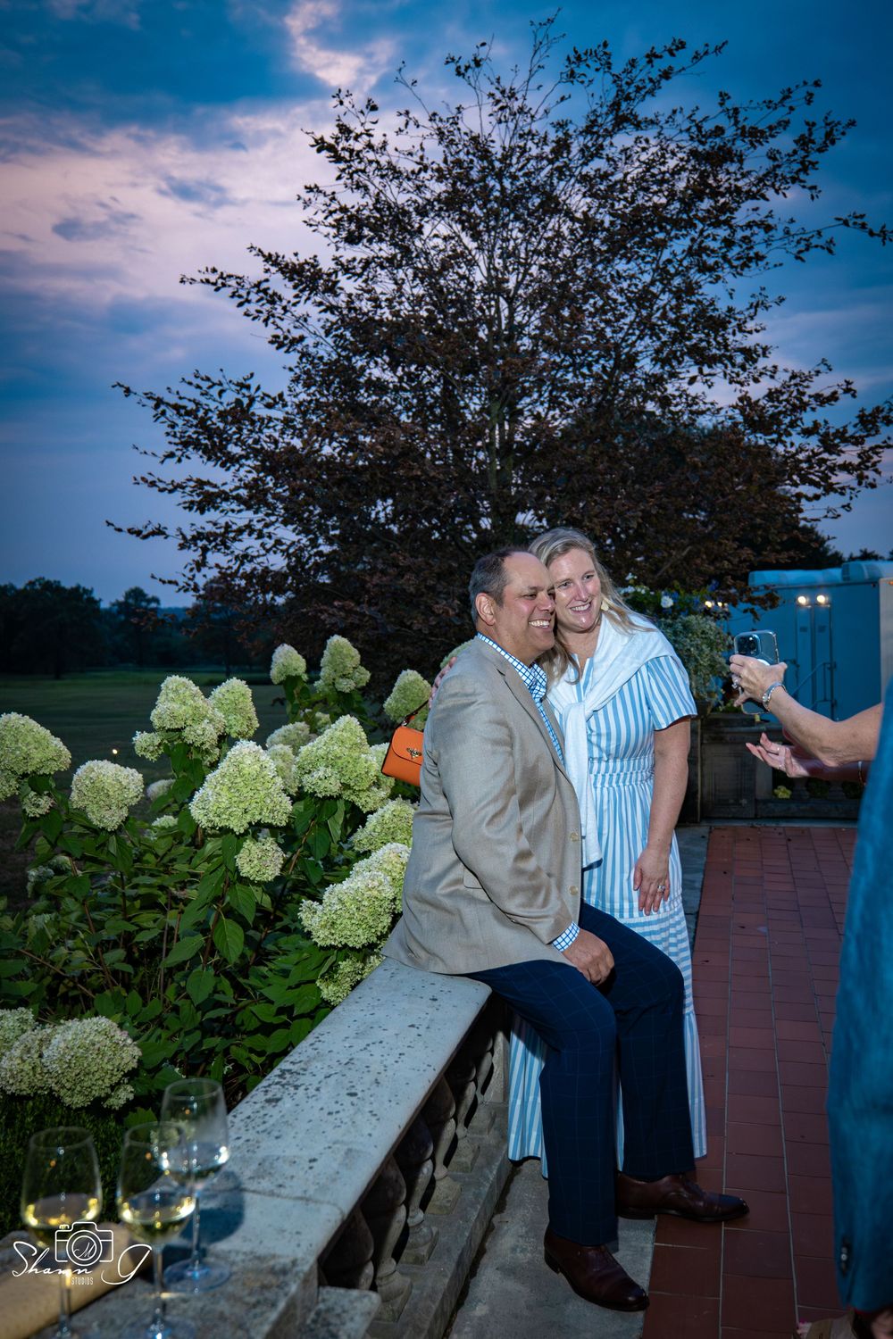 Couple posing together on a terrace at dusk, surrounded by white hydrangeas and lush green trees.