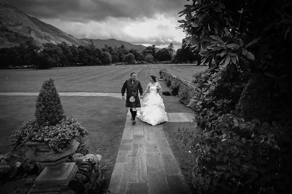Black and white photo of a bride and groom walking hand in hand through elegant garden grounds with misty mountains behind.