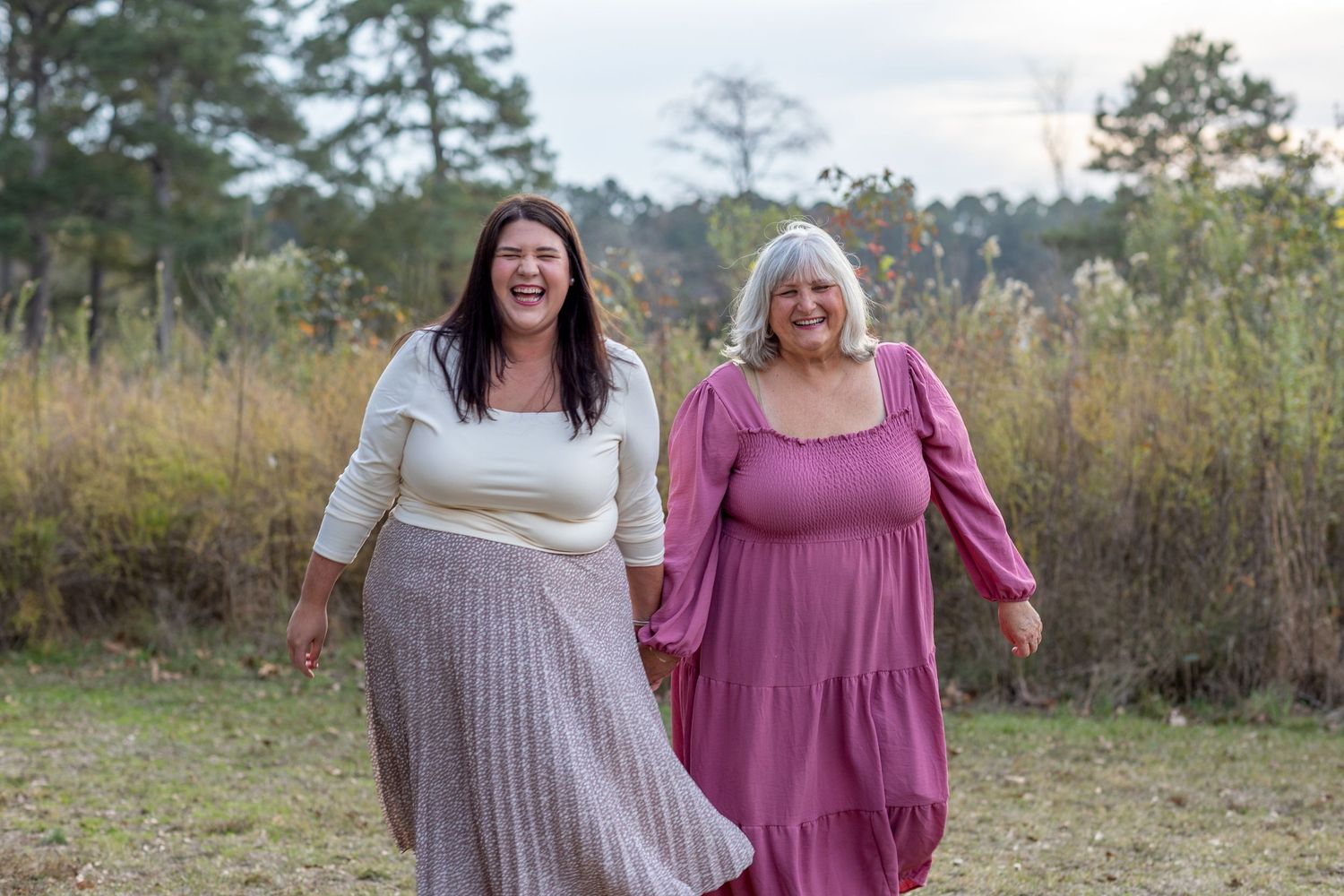 Two women smiling outdoors in elegant dresses, one in white and gray, one in pink, standing in a natural setting.
