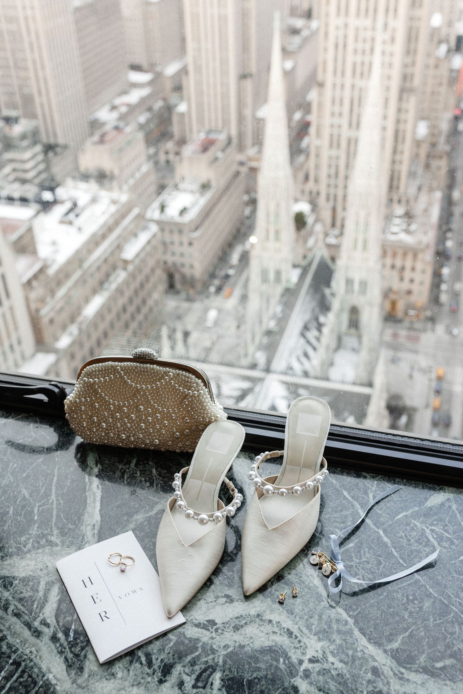 Bridal heels with pearl details, clutch purse, and jewelry displayed on marble surface overlooking NYC skyline.