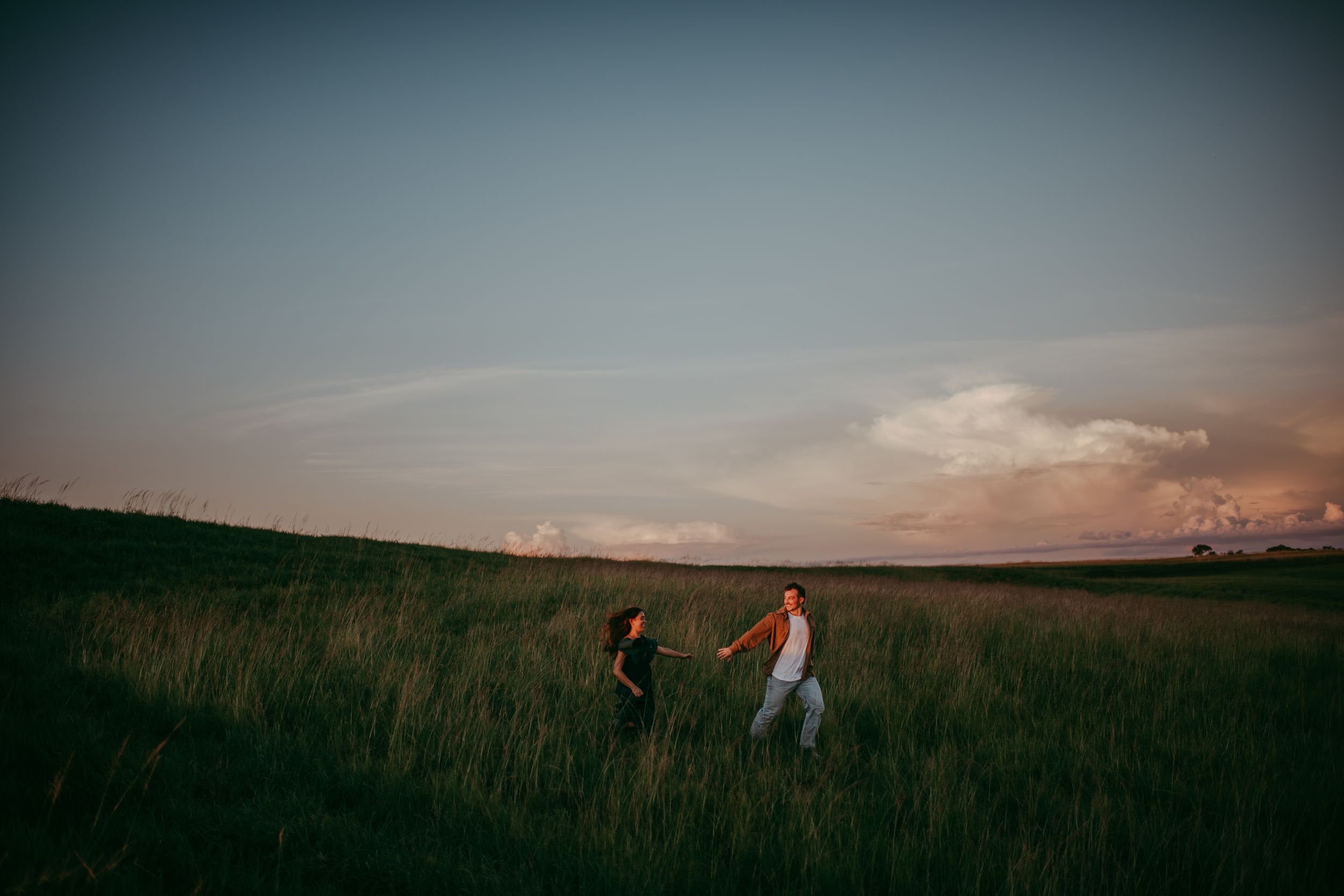 Couple holding hands walking through tall green grass field at sunset with dramatic pink clouds in the sky.