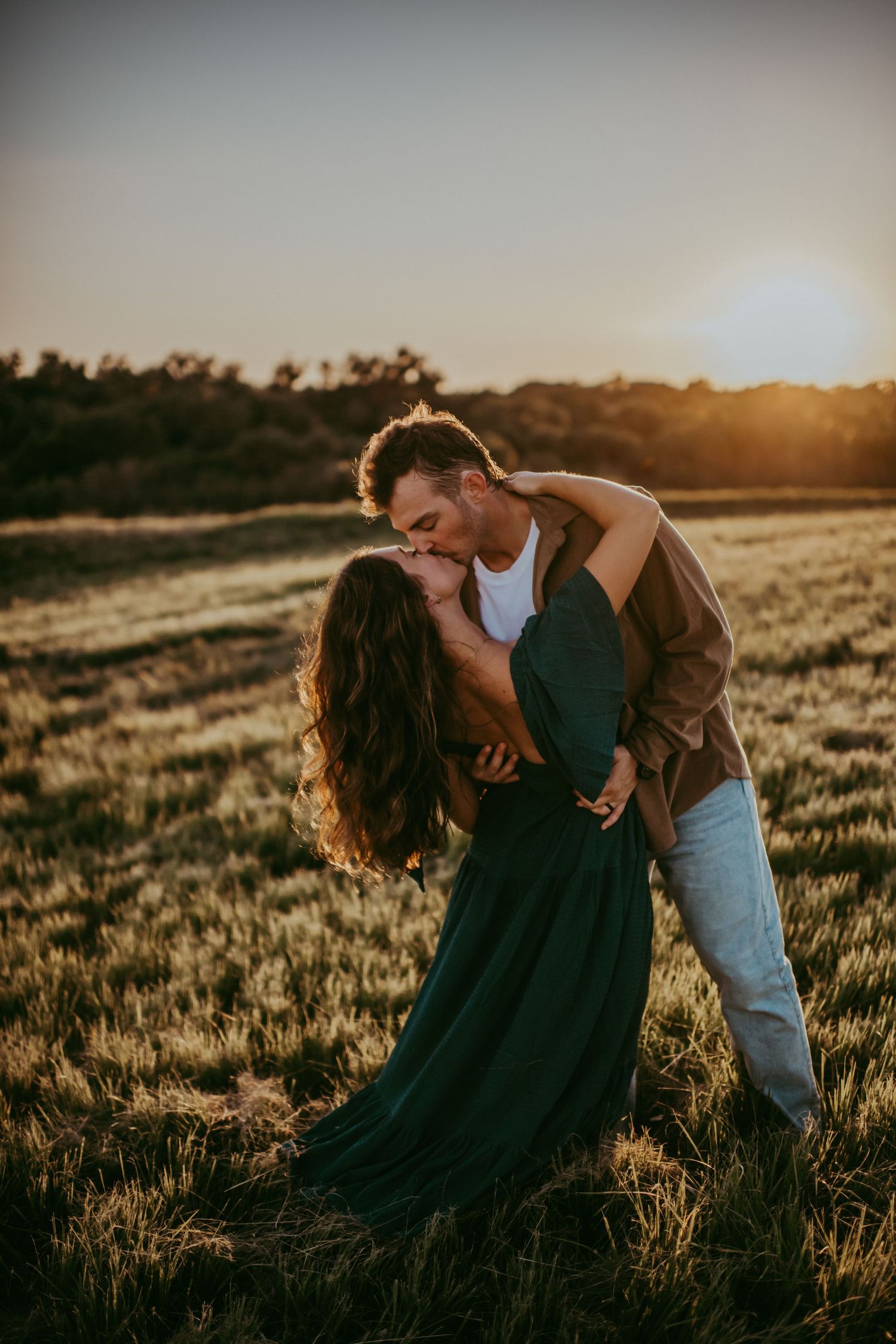 Couple sharing a romantic dip kiss in a golden field at sunset, woman in teal dress, man in brown jacket.