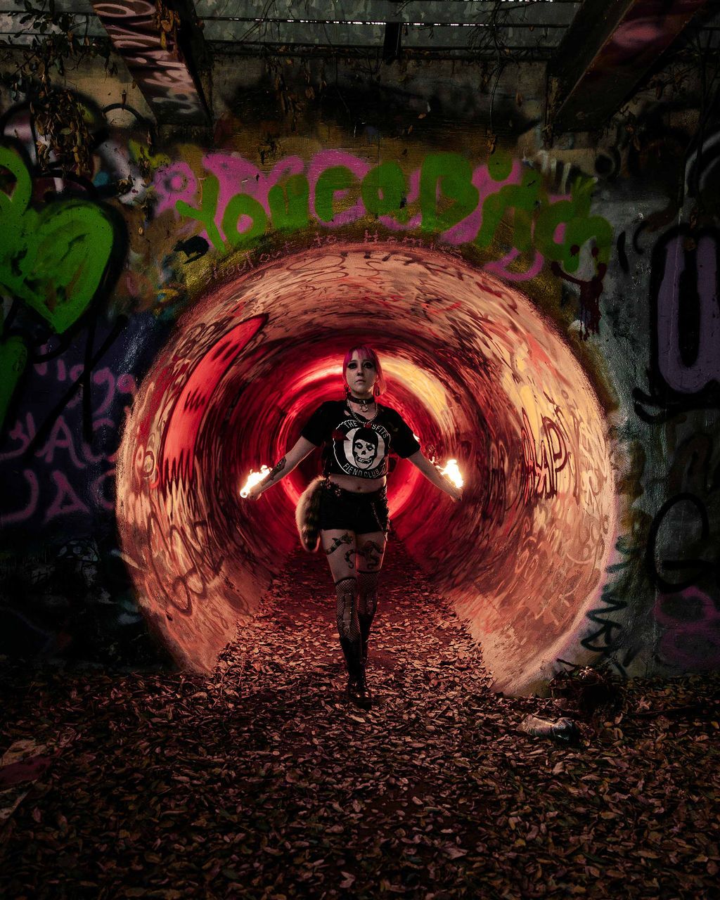 a female fire performer emerges from a large graffiti-covered pipe with theatrical lighting