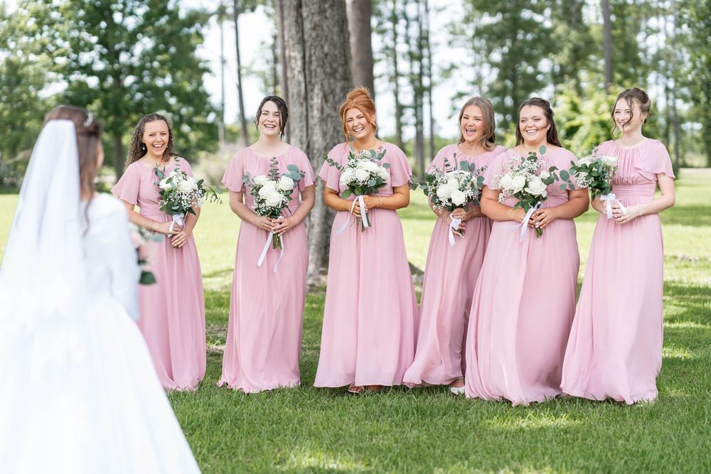Bride in white gown faces six bridesmaids in pink floor-length dresses holding white bouquets on a green lawn.