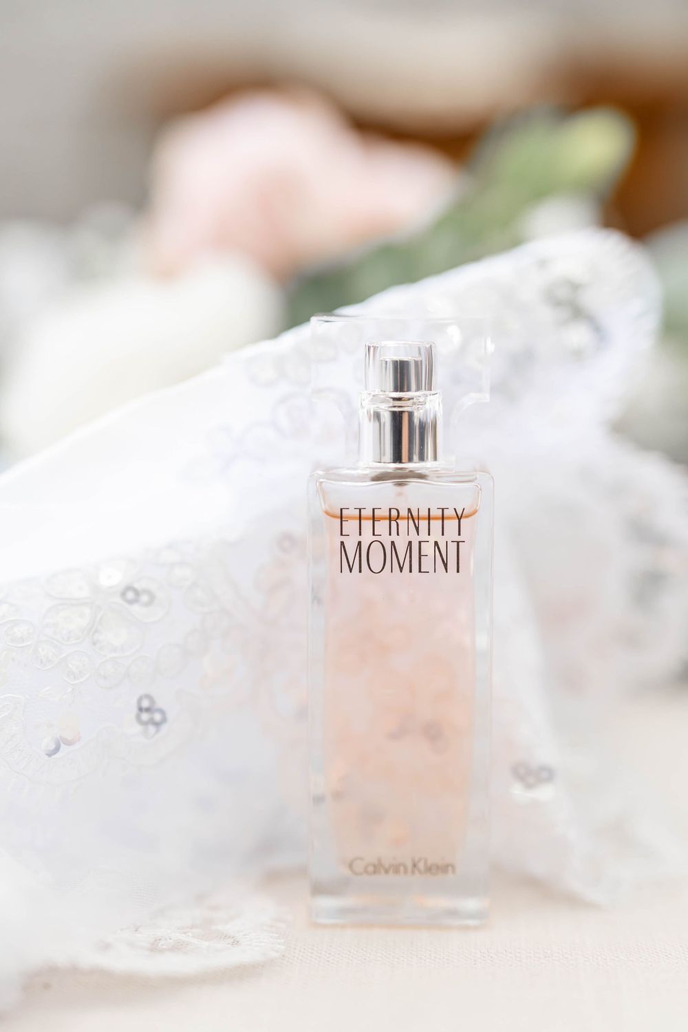 Calvin Klein Eternity Moment perfume bottle resting on white lace fabric with soft floral bokeh background.