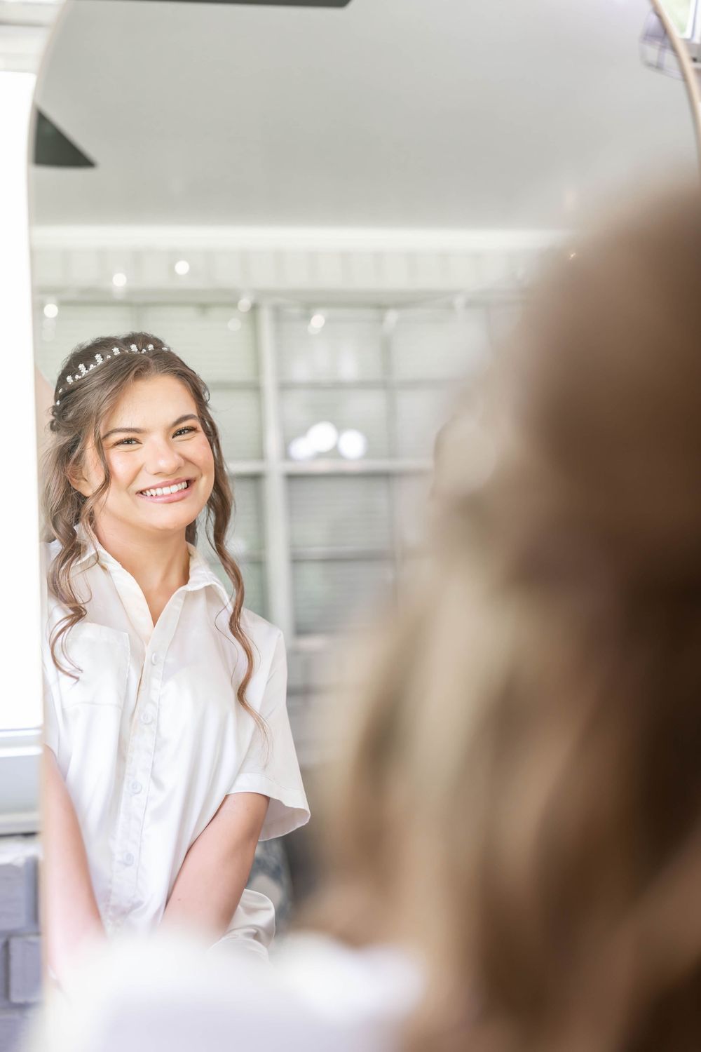 Smiling bride in white shirt with floral hair accessory admiring herself in mirror while getting ready.