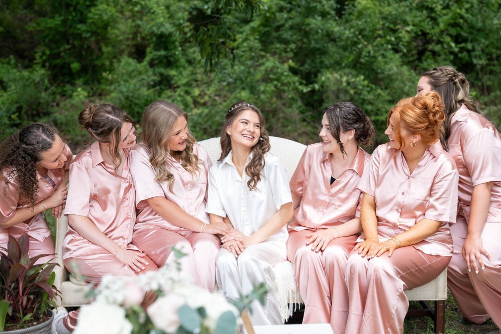 Bride in white robe laughing with bridesmaids in pink satin pajamas outdoors before wedding ceremony.