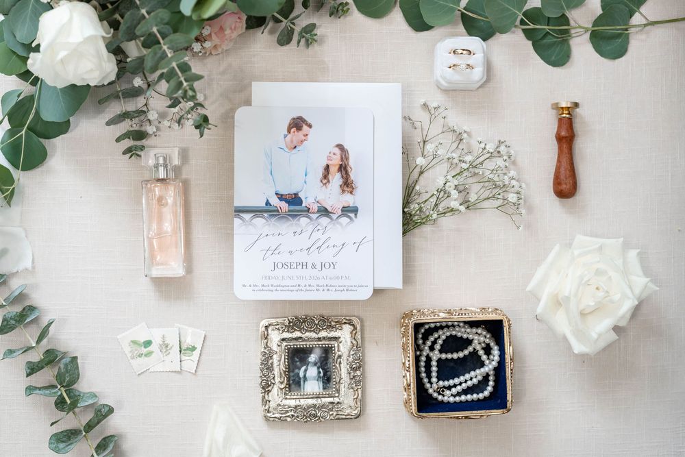 Elegant wedding flat lay with invitation, pearl bracelet, rings, white rose, eucalyptus, and perfume on linen background.
