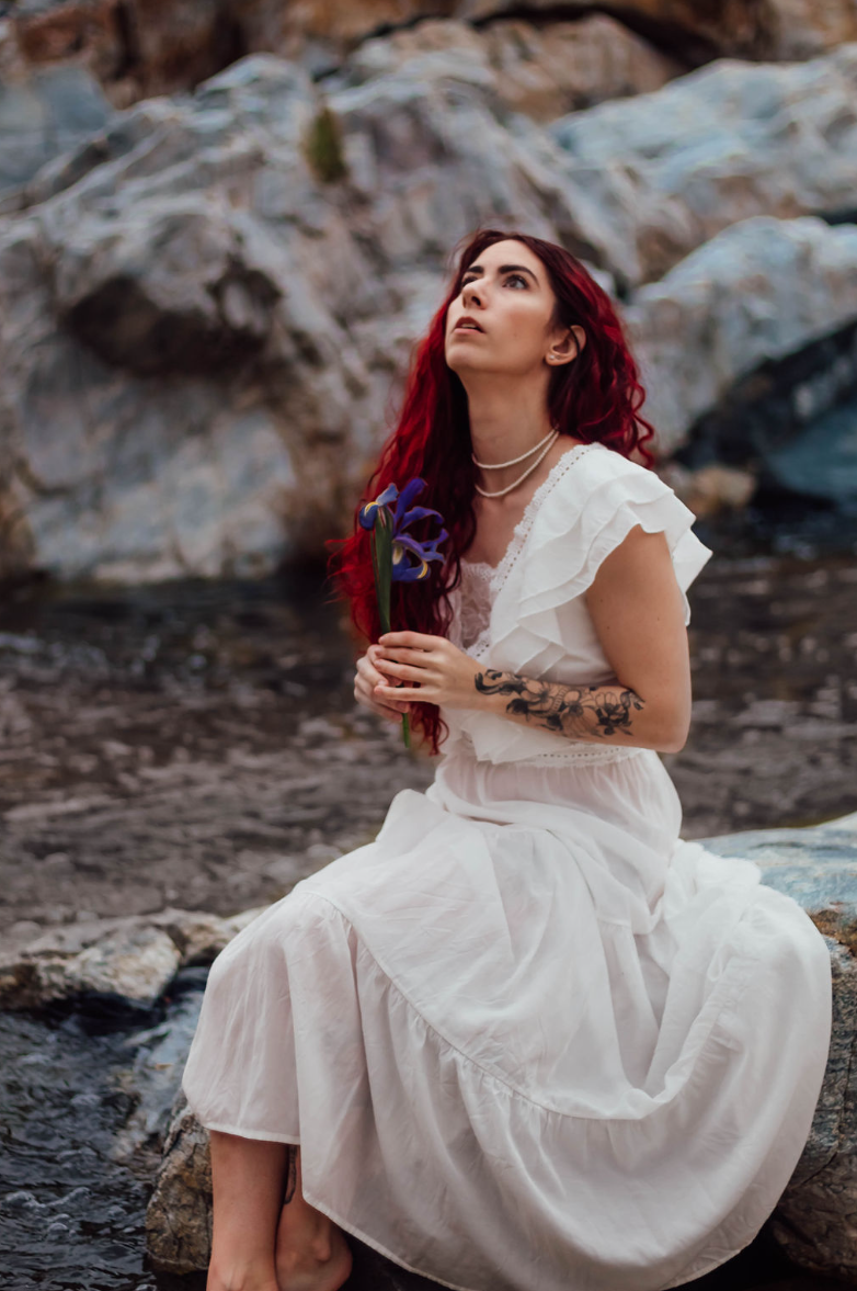 Redhead woman sits at a waterfall holding a flower and staring up at the sky in a portrait photography session.