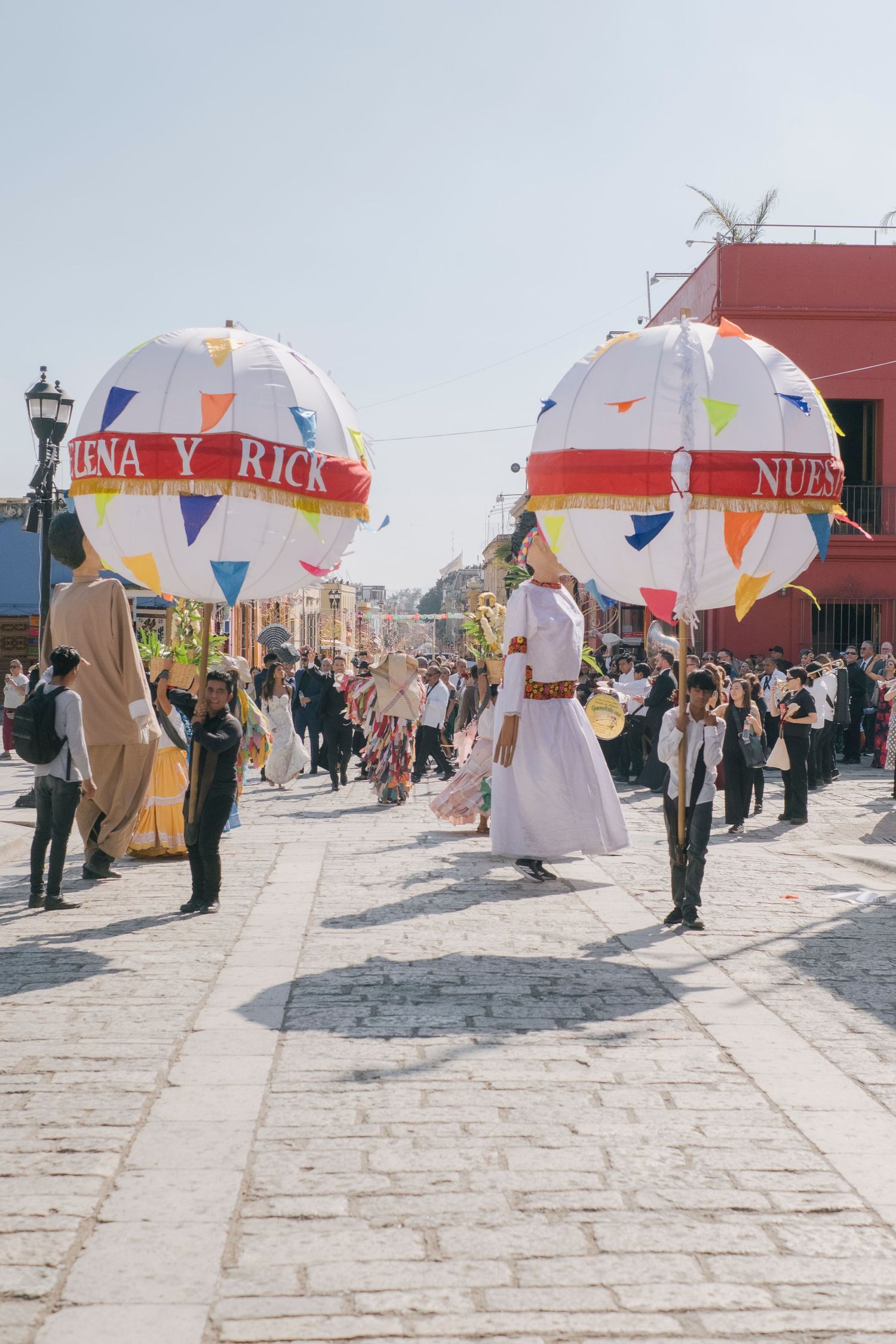 Colorful Mexican street parade with participants carrying large decorative paper balloons on a sunny cobblestone street.