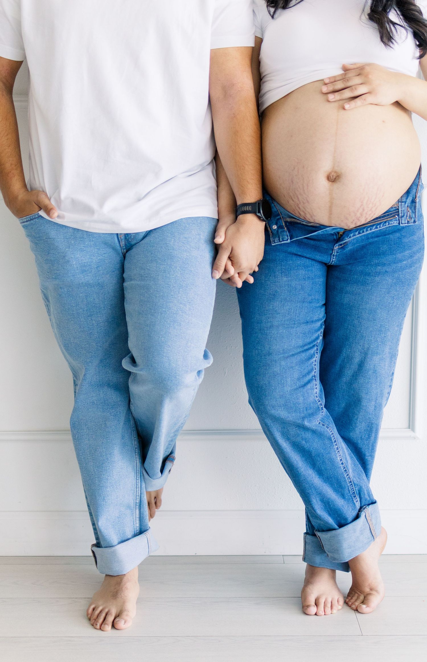 Detail maternity image of mom and dad, holding hands and leaning against a white wall in a natural light studio