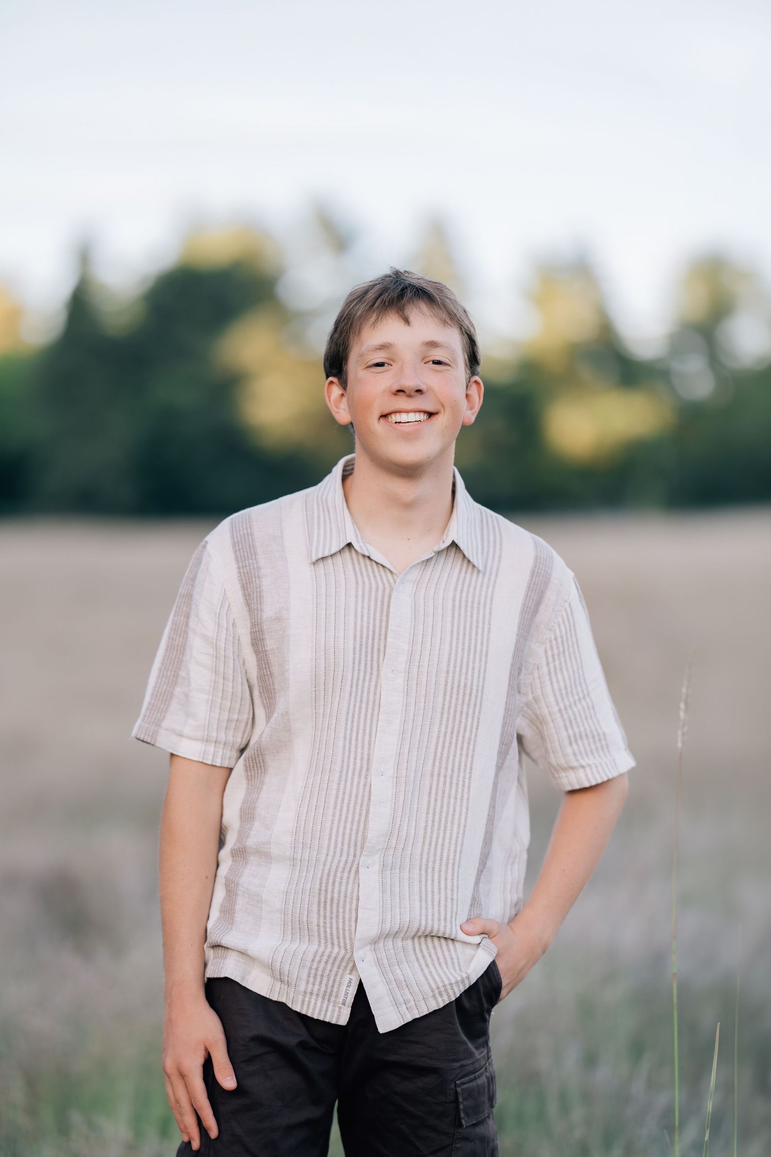 Smiling teenage boy in striped linen shirt standing in an open field with trees in the background.