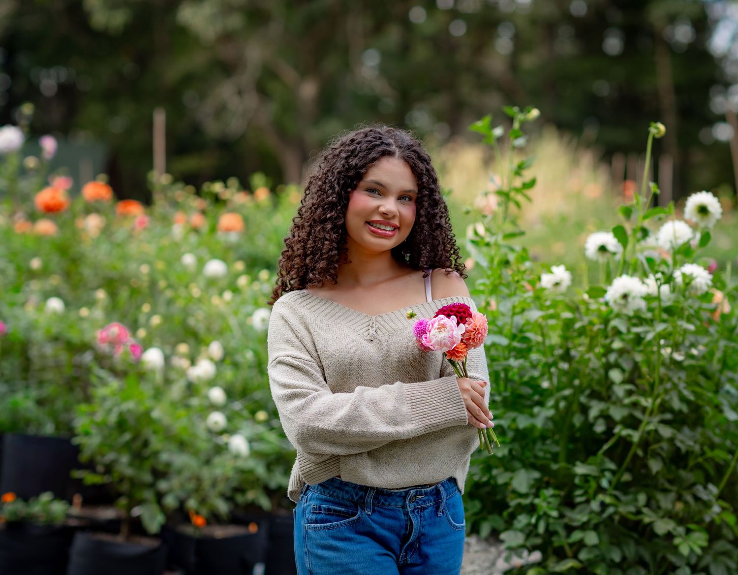 Young woman with curly hair holding pink flowers, smiling in a lush garden with colorful dahlias blooming behind her.