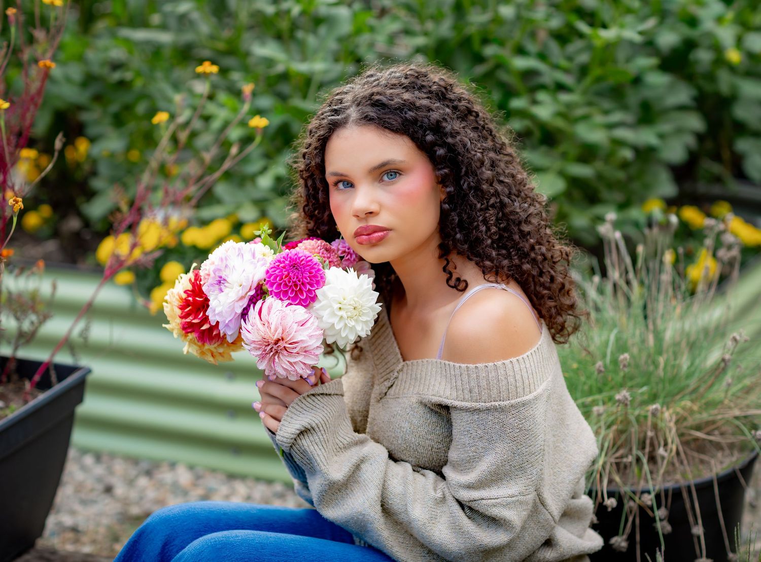 Young woman with curly hair holds pink and white dahlias in a garden, wearing an off-shoulder gray sweater.