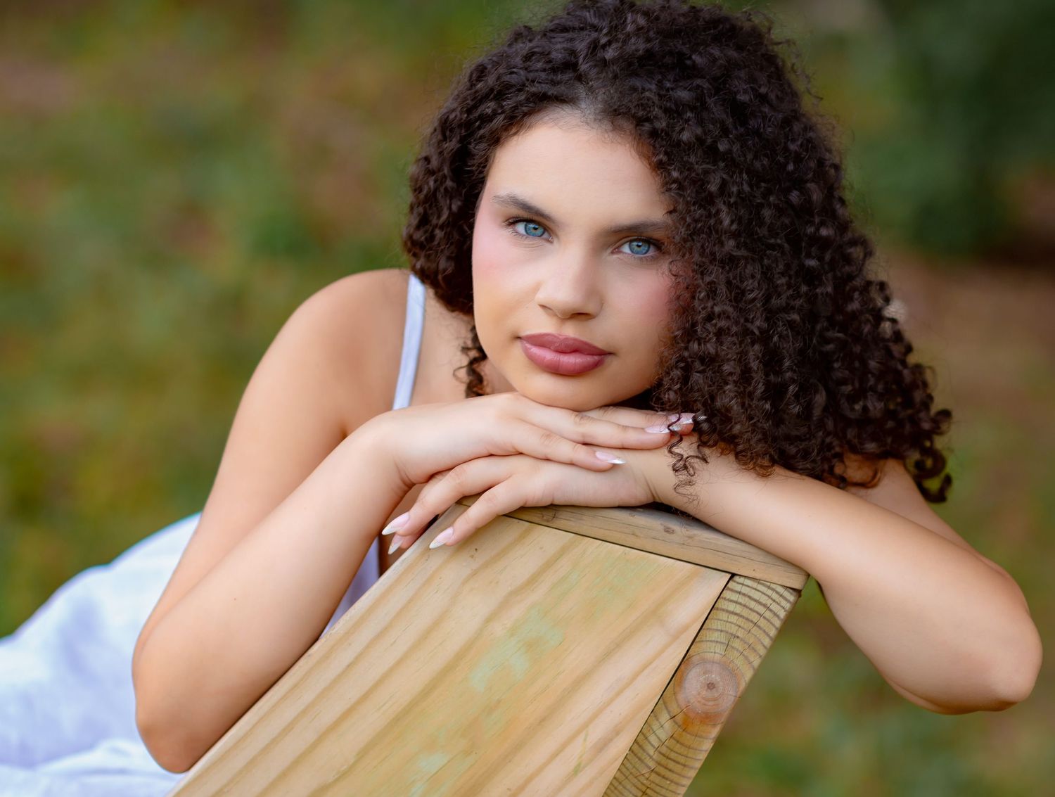 Young woman with curly hair resting her chin on hands atop a wooden fence outdoors.