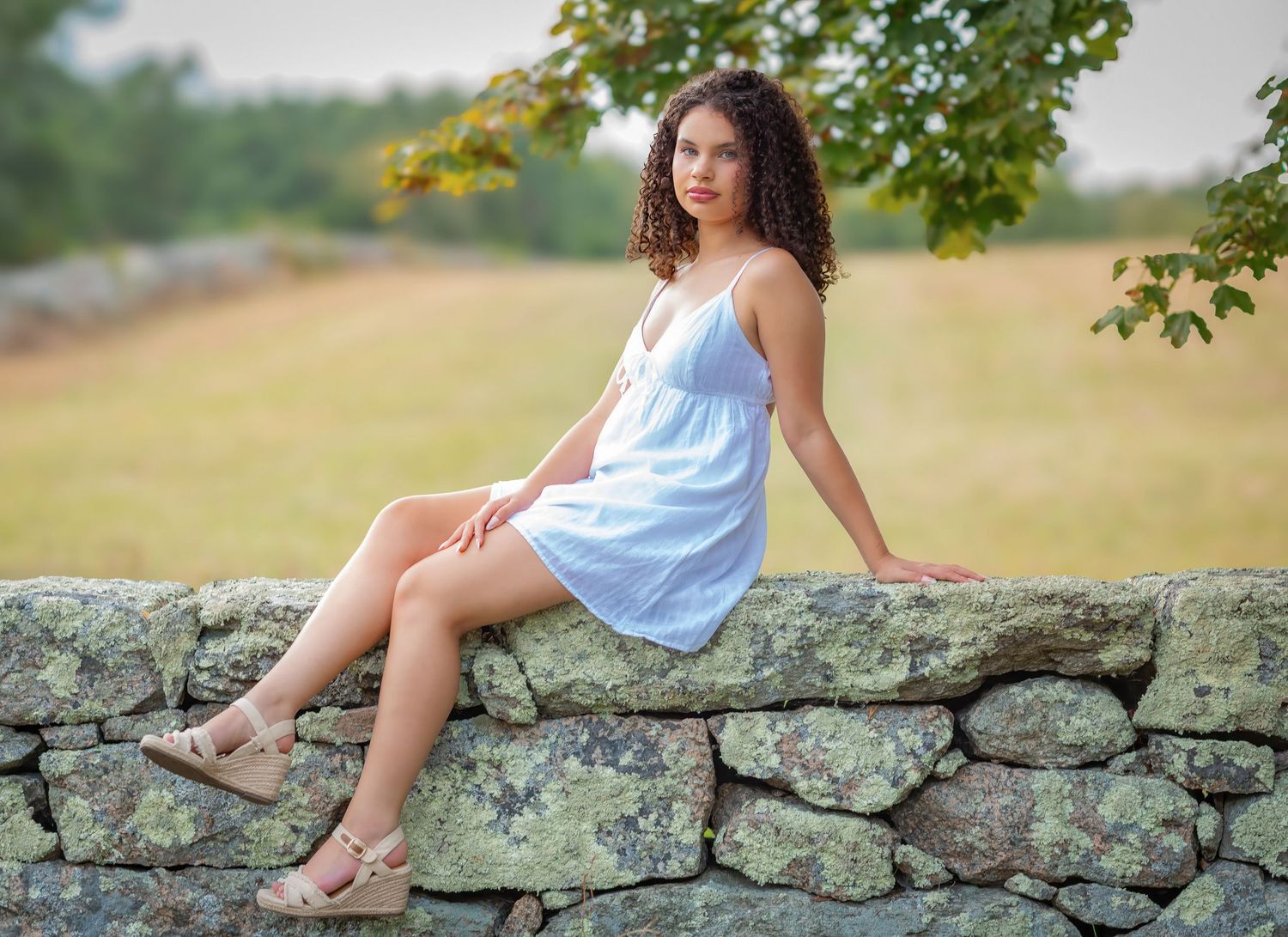 Young woman with curly hair in a light blue sundress sitting on a stone wall in a scenic outdoor setting.