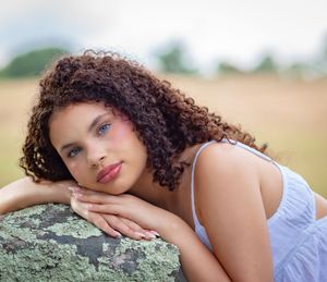 Young woman with curly hair and blue eyes resting on a mossy rock in a serene outdoor field setting.