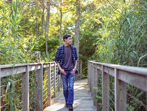 Young man in plaid shirt walking on wooden boardwalk surrounded by lush green trees and tall grass.