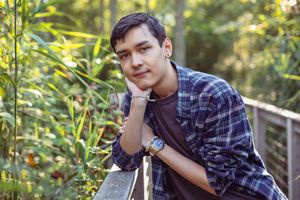 Young Asian man in plaid shirt resting chin on hand, sitting outdoors surrounded by lush green foliage.