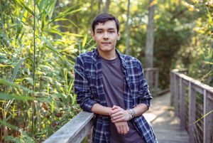 Young man in plaid shirt leaning on wooden bridge railing surrounded by lush green foliage outdoors.