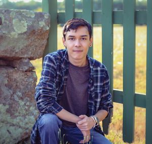 Young man in plaid shirt sitting by stone wall and green fence outdoors, smiling casually.