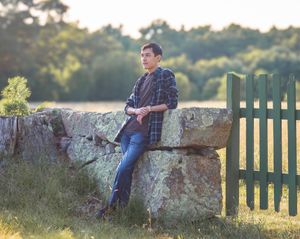 Young man in plaid shirt and jeans leaning against a stone wall near a green gate in a sunny field.