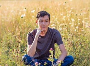 Young man sitting cross-legged in a sunlit wildflower meadow, resting chin on hand in a thoughtful pose.